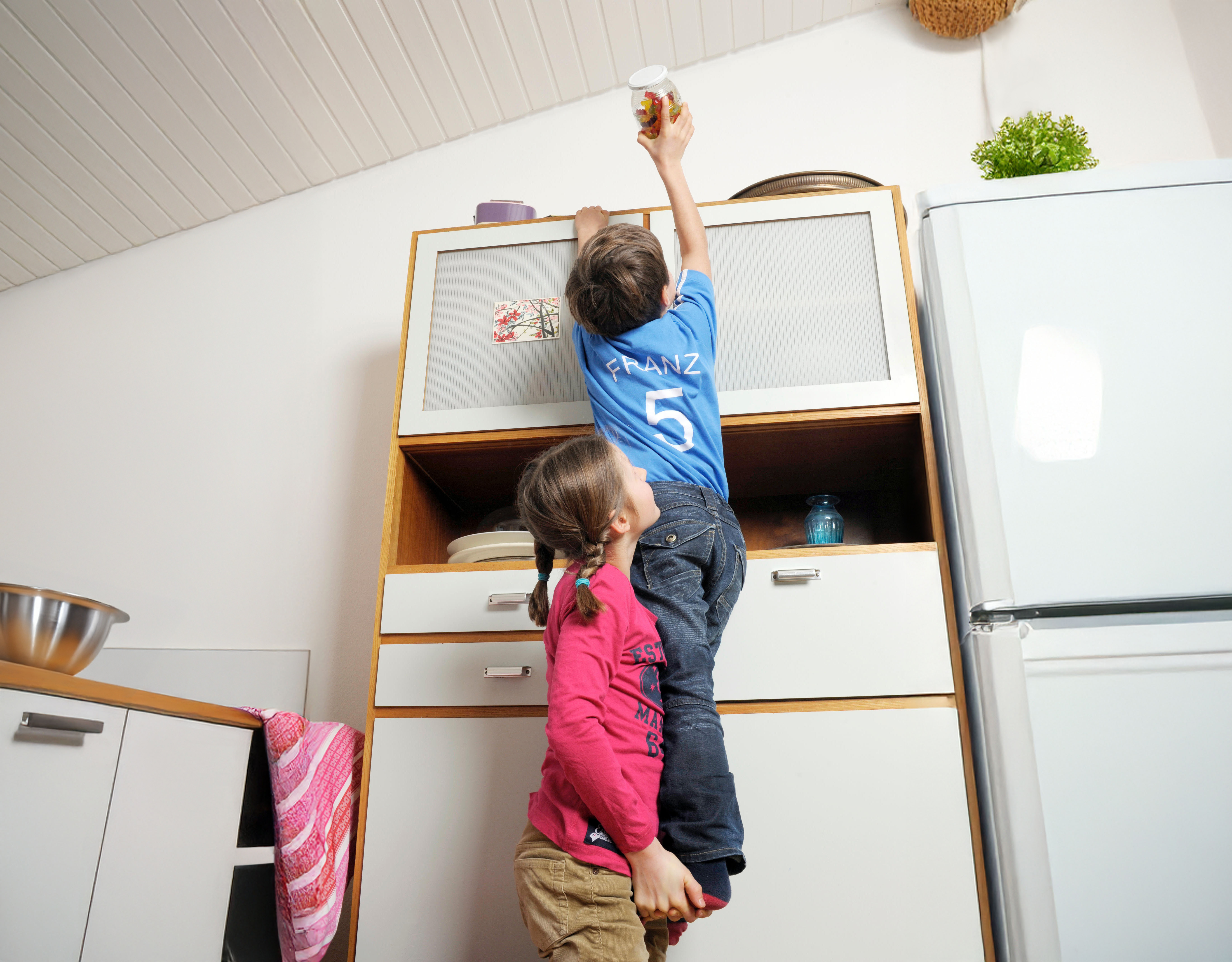 Ein Kind hebt ein anderes mit Räuberleiter, um Süssigkeiten auf dem Küchenschrank zu erreichen.